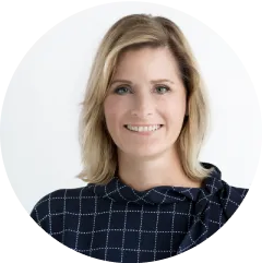 Eine Frau mit blondem Haar, die ein marineblaues Oberteil mit weißem Gittermuster trägt, lächelt vor einem schlichten weißen Hintergrund in die Kamera.